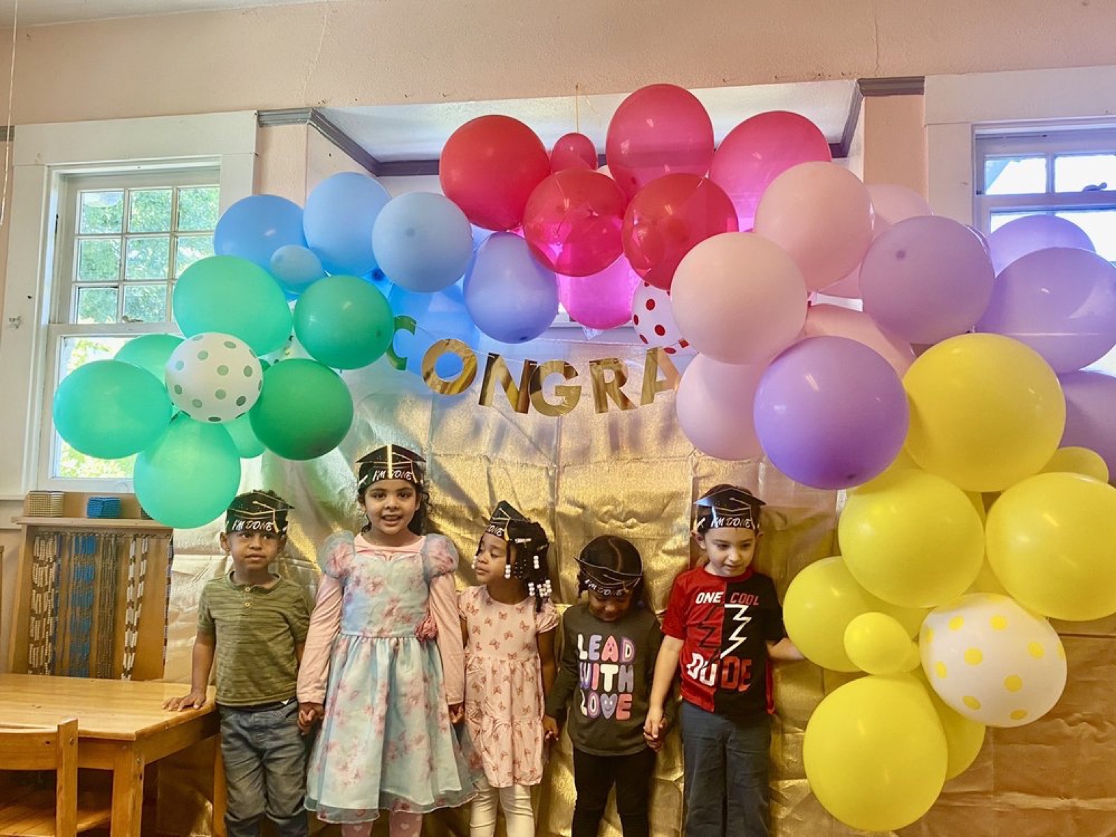 A group of young graduates in caps and gowns celebrating beneath the CONGRATS banner and balloon arch