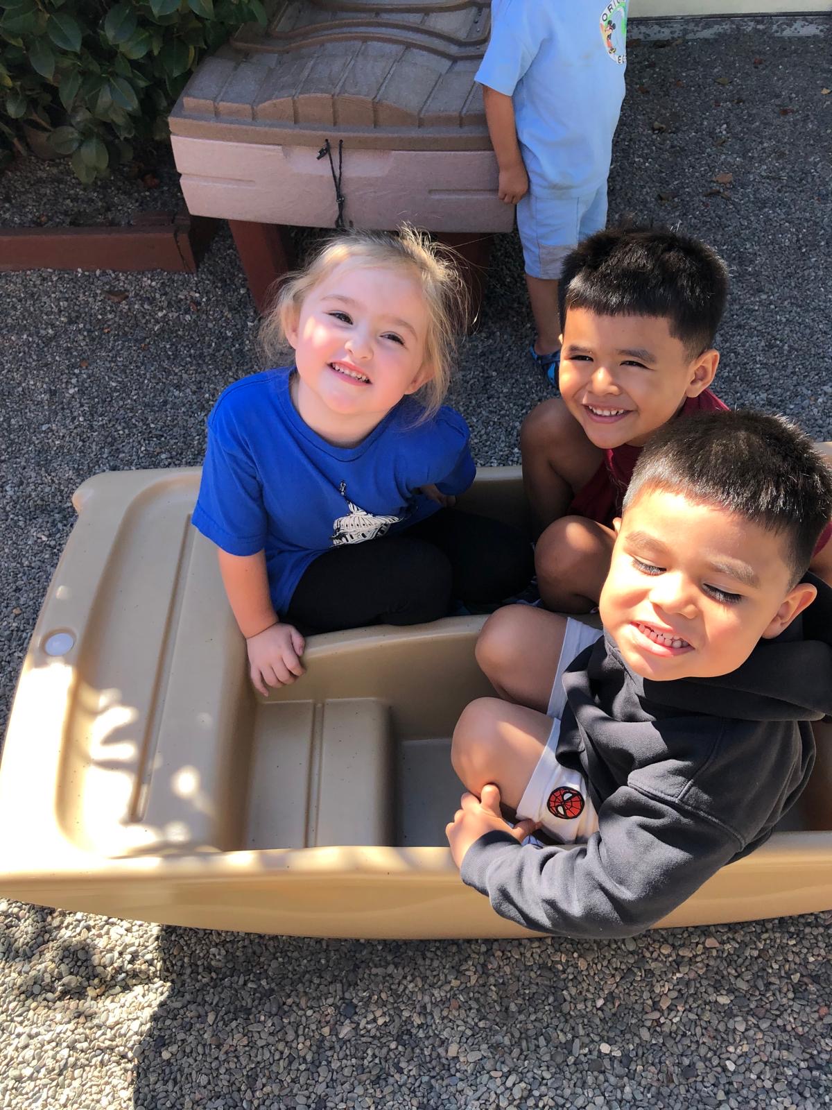 Three children grin from inside a play boat in the sunshine