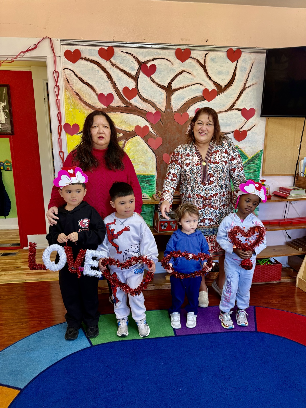 Teachers and children hold tinsel hearts and the word LOVE before the heart-covered tree mural